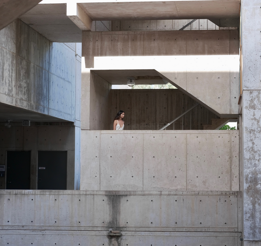 A woman in a white slip dress stands on a concrete balcony within a brutalist architectural structure. The geometric composition of the space features raw concrete walls, angular staircases, and intersecting planes, creating a sense of depth and layering. Harsh sunlight casts strong shadows, emphasizing the texture and modularity of the building. The woman's delicate presence contrasts with the rigid, industrial aesthetic of the environment.