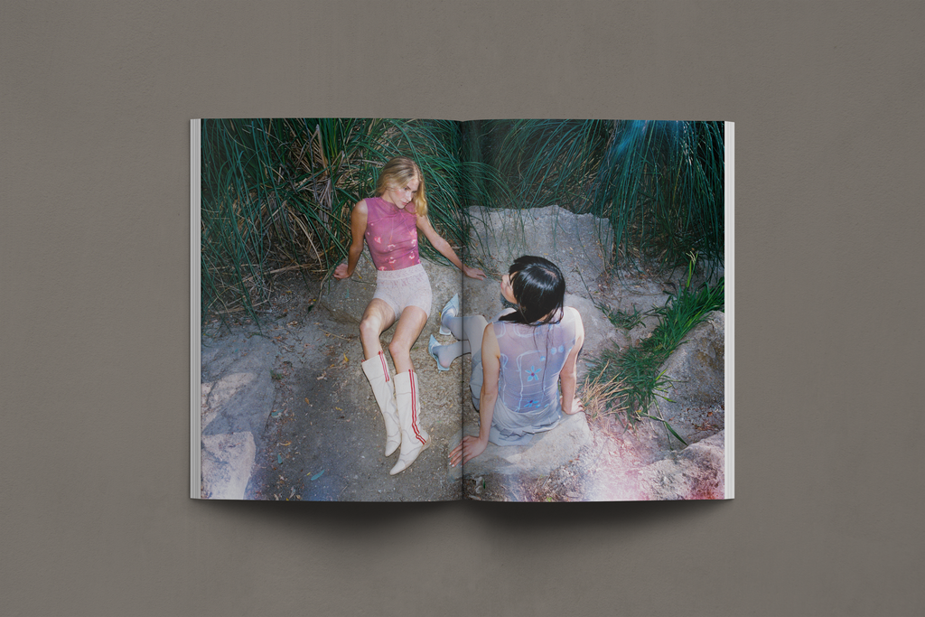 Two models pose in a lush, natural setting, wearing sheer printed tops that blend with the organic surroundings. One model raises her arm gracefully, while the other gazes downward, her hair adorned with a delicate feather. The soft translucency of the fabrics interacts with the light, creating an ethereal, dreamlike atmosphere that evokes themes of harmony, movement, and connection to nature.