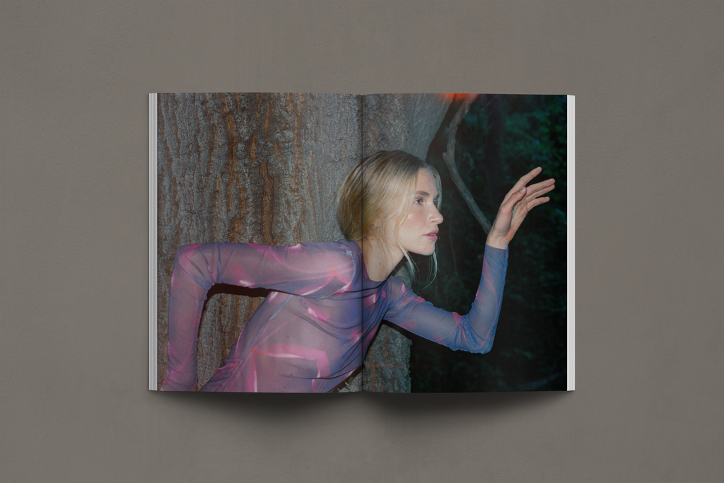 This editorial spread showcases a model in a sheer, printed long-sleeve top with abstract organic patterns. The deep forest setting and interplay of light and shadow enhance the ethereal quality of the composition. Her pose, with an outstretched hand and gaze into the distance, evokes a sense of movement and connection with the environment. The contrast between her luminous skin and the dark natural backdrop adds to the mysterious and dreamlike atmosphere.