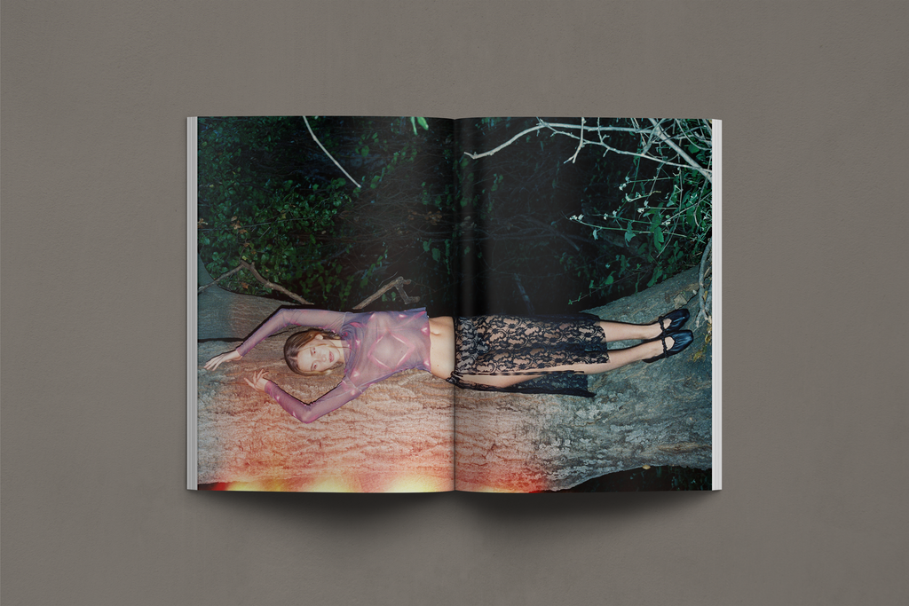This editorial image features a sheer printed top and a black lace skirt, blending delicate transparency with intricate texture. The model is sprawled across a fallen tree, evoking themes of raw nature and ethereal beauty. The atmospheric lighting and deep forest setting create an otherworldly mood, enhancing the dreamlike quality of the composition. The contrast between soft fabric and organic elements highlights the interplay between fashion and the natural world.