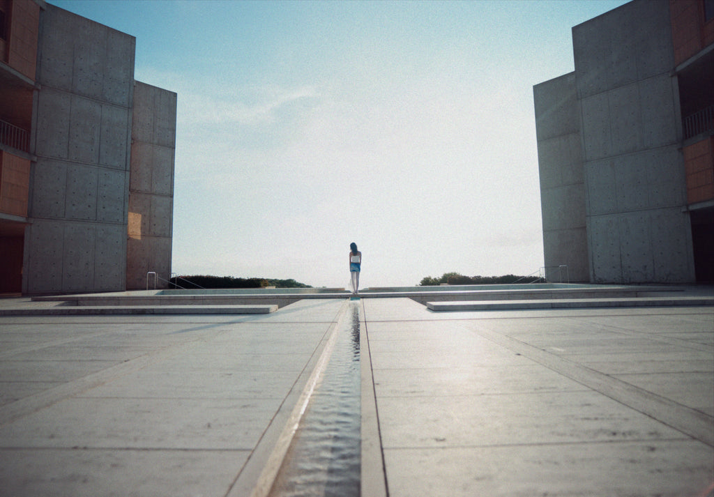 captures a lone figure walking along the central water feature of the Salk Institute in La Jolla, California. The brutalist concrete buildings frame the composition, leading toward the horizon where the ocean meets the sky. The vast open space, soft lighting, and minimalist architecture create a serene, meditative atmosphere.