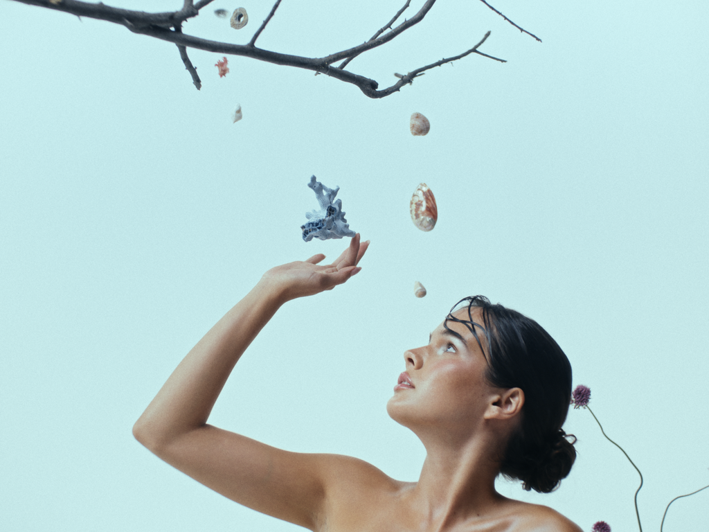  a woman with slicked-back hair gazing upwards, her hand delicately raised toward floating seashells, coral, and organic forms suspended in midair. A bare tree branch extends into the frame, adding an ethereal, surreal quality. The soft blue-toned lighting creates a dreamlike atmosphere, emphasizing the connection between nature, the ocean, and the human form.