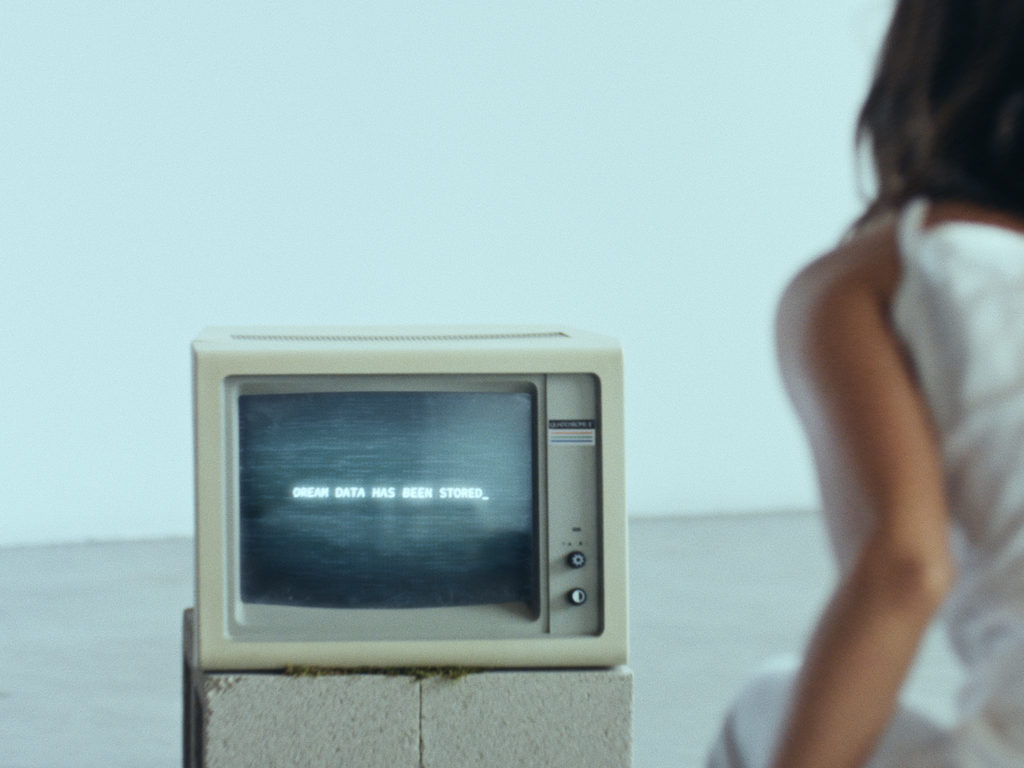 A vintage computer monitor with a grainy black-and-white screen displays the text "DREAM DATA HAS BEEN STORED." The monitor has an old-school aesthetic with knobs and buttons on the side. Partially visible on the right is a person with dark hair wearing a light-colored garment, adding a mysterious element. The background is a minimalist white space, enhancing the futuristic yet nostalgic atmosphere.