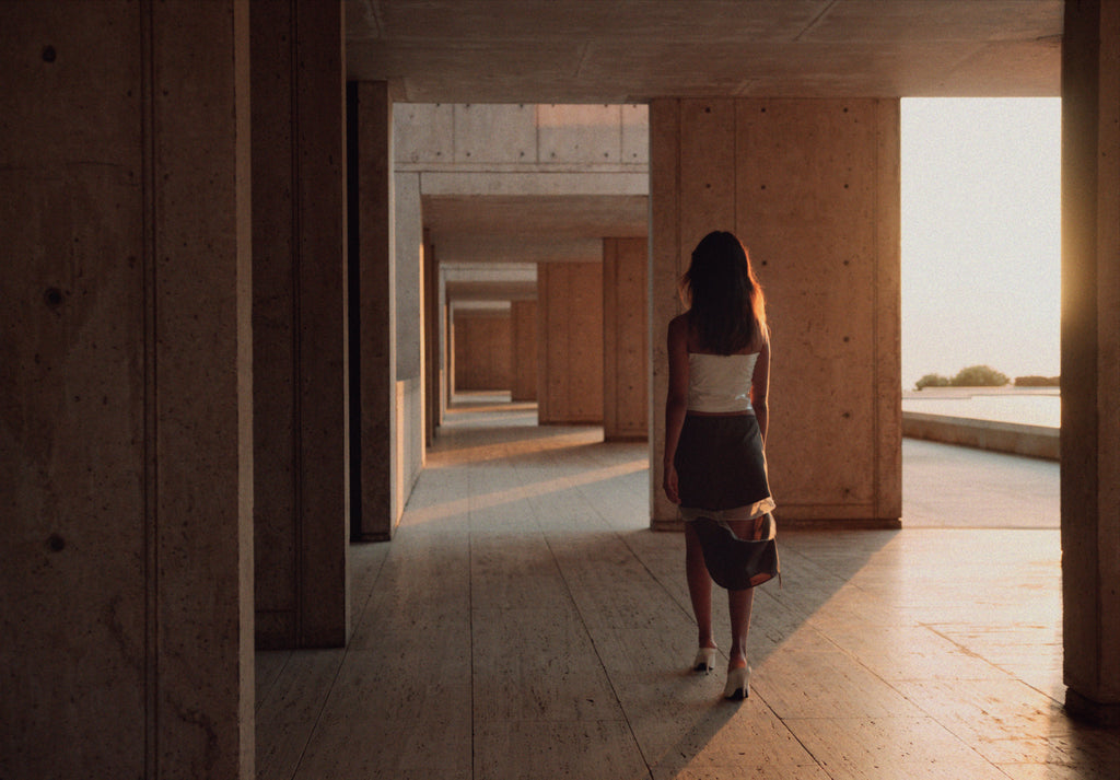 a woman walking through the concrete corridors of the Salk Institute at sunset. She wears a white sleeveless top and a dark skirt with a sheer, flowing underlayer, paired with white heels. The golden light casts elongated shadows on the travertine floor, emphasizing the geometric repetition of the brutalist architecture. The open corridor leads to an expansive ocean view, blending the structure seamlessly with the natural horizon