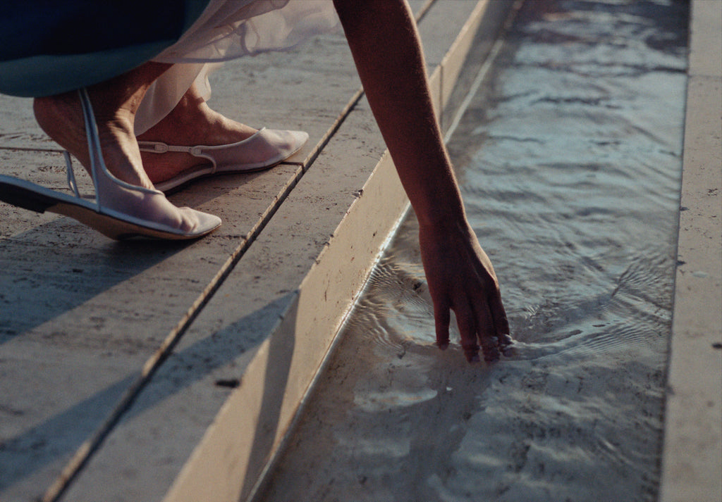 Close-up of a model’s hand touching the flowing water feature at the Salk Institute, with sheer silk fabric and delicate white slingback flats. A serene moment blending minimalist fashion with architectural elegance.