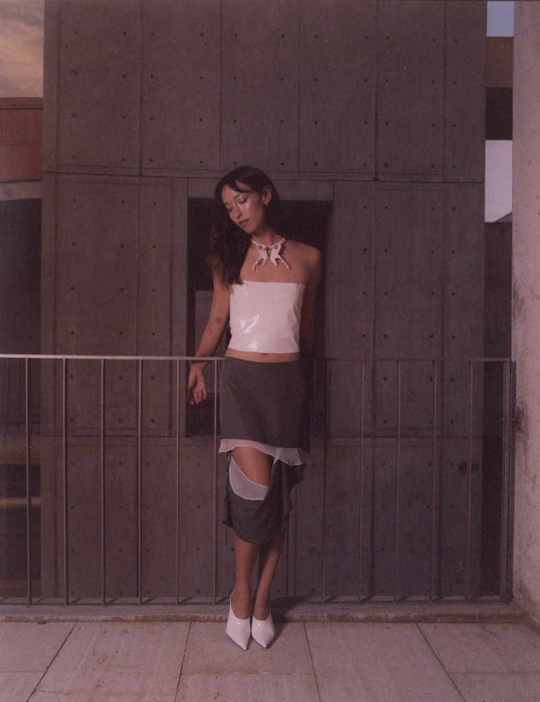 Model wearing a minimalist white silk tube top, deconstructed dark grey asymmetrical skirt with sheer layers, and sculptural statement necklace. Avant-garde sustainable fashion shot at the Salk Institute, blending modern architecture with contemporary womenswear.