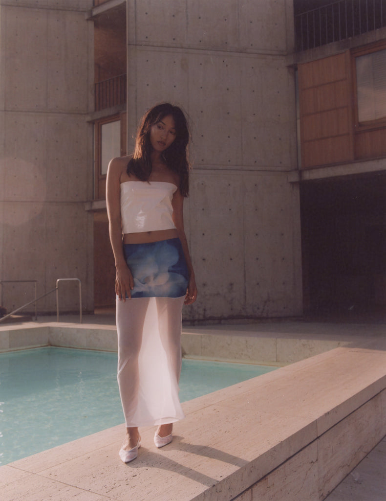Model wearing a silk skirt with a blue floral digital print and a sheer white overlay, standing near a pool at the Salk Institute. The skirt's fluid design is inspired by nature and architecture, reflecting soft, ethereal movement.