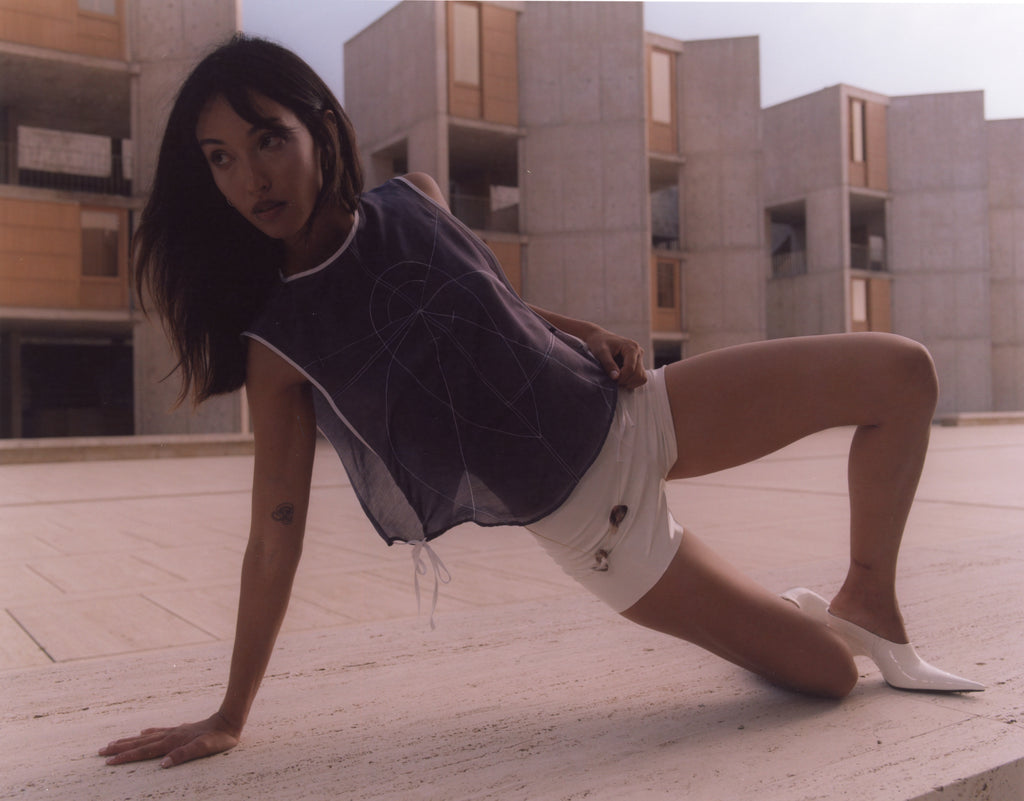 Model wearing a sheer navy silk top with embroidered sacred geometry, paired with white patent shorts and pointed mules. Captured in dynamic motion at the Salk Institute, merging contemporary fashion with brutalist architecture.