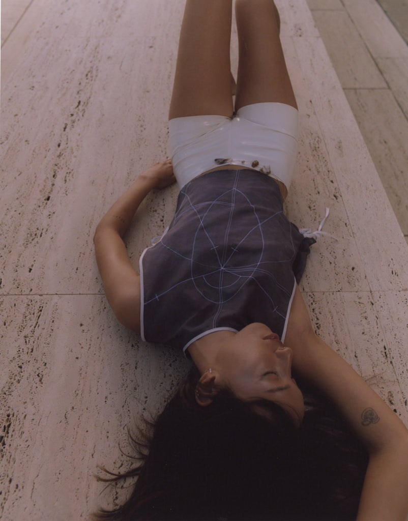 Model wearing a dark blue silk top with intricate geometric embroidery, inspired by sacred geometry, paired with high-shine white patent shorts. Photographed lying on travertine stone at the Salk Institute, blending fashion with architectural minimalism.