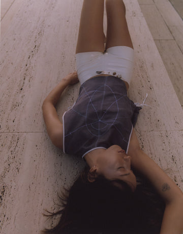 Model wearing a dark blue silk top with intricate geometric embroidery, inspired by sacred geometry, paired with high-shine white patent shorts. Photographed lying on travertine stone at the Salk Institute, blending fashion with architectural minimalism.